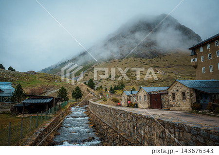 Mountain stream flowing between stone walls past old stone buildings below a fog covered peak in Vall de Nuria 143676343