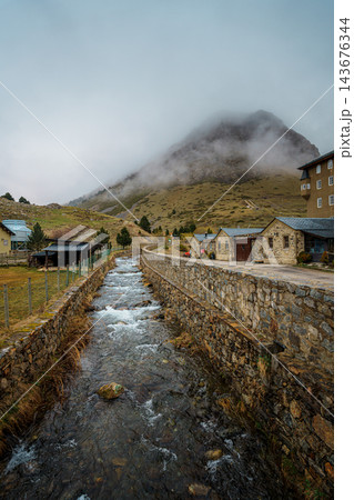 Mountain stream flowing between stone walls past old stone buildings below a fog covered peak in Vall de Nuria Mountain stream flowing between stone walls past old stone buildings below a fog covered peak in Vall de Nuria 143676344