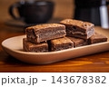 Stacked Homemade Chocolate Brownies on a Plate with Coffee Cup Close-Up in Cozy Cafe Setting 143678382