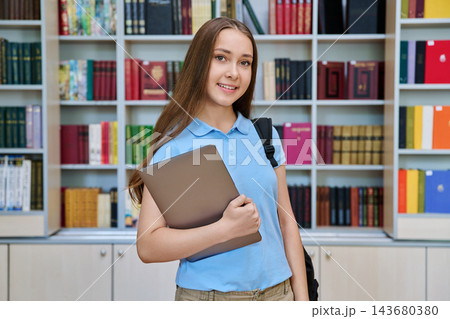 Teenage girl student holding laptop with backpack in library portrait 143680380