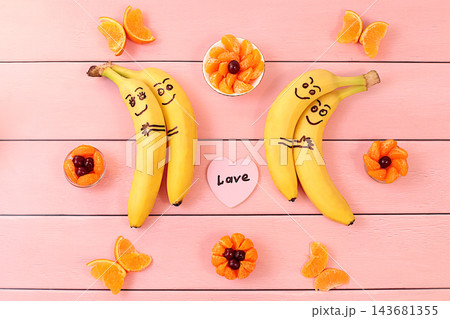 Adorable bananas in love, hugging each other, accompanied by a small heart labeled love and surrounded by butterflies and flowers made from mandarin oranges. This playful treat is perfect for children Adorable bananas in love, hugging each other, accompanied by a small heart labeled love and surrounded by butterflies and flowers made from mandarin oranges. This playful treat is perfect for children 143681355