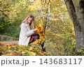 Young woman enjoys a colorful autumn walk in a city park, making a maple leaf wreath and playfully throwing fallen leaves. Seasonal lifestyle, outdoor recreation, wellness and the beauty of fall 143683117