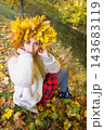 Young woman wearing a wreath of colorful maple leaves relaxes in an autumn park. Seasonal lifestyle portrait celebrating fall colors, outdoor wellness and the beauty of nature. 143683119