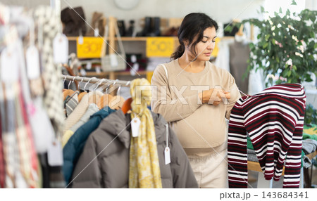 Pregnant Latin girl is busy looking at clothes items in store, choose sweater 143684341