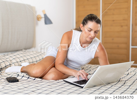 Pretty woman in nightgown works on laptop sitting on a bed in bedroom 143684370