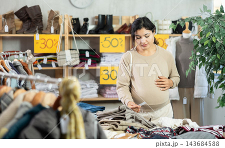 Pregnant mulatto young female client choosing trousers in clothing mall 143684588
