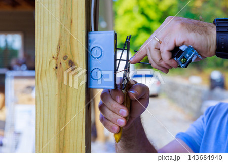 An electrician connects wires in switch box mounted on wooden post at an outdoor site 143684940