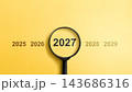 Magnifying glass focusing on the year 2027 among sequential years on a yellow background, representing future planning, time management, and business forecasting. Concept of Happy New Year 143686316