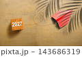 Red paper airplane flying over sand background with number 2027 on wooden cube and palm tree shadow. Concept of Happy New Year 143686319