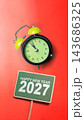 Happy New Year 2027 message on a green sign with a black and lime green alarm clock pointing almost to midnight, set against a solid bright red background. Concept of Happy New Year 143686325