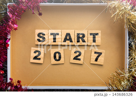 Start 2027 spelled out with wooden letter and number tiles inside a festive box decorated with pink and gold tinsel, top-down view. Concept of Happy New Year 143686326