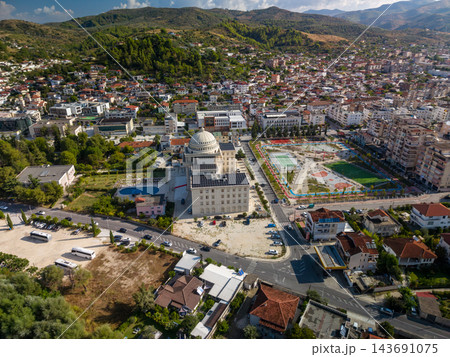 Berat Albania fills broad valley beneath wooded mountains, seen from above. City buildings and red roof houses cluster around prominent domed structure and colorful outdoor courts. Berat Albania fills broad valley beneath wooded mountains, seen from above. City buildings and red roof houses cluster around prominent domed structure and colorful outdoor courts. 143691075