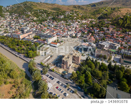 Berat, Albania appears in sunlit aerial view, with hillside neighborhoods surrounding broad paved plaza. River, tree lined roads and tightly packed buildings define town setting, amid green hills 143691078