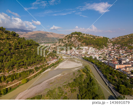 Berat Albania rests along Osum River, with dense pale buildings beneath forested ridge. Gravel riverbanks, surrounding hills and blue sky give historic town broad scenic setting, with rooftops Berat Albania rests along Osum River, with dense pale buildings beneath forested ridge. Gravel riverbanks, surrounding hills and blue sky give historic town broad scenic setting, with rooftops 143691079