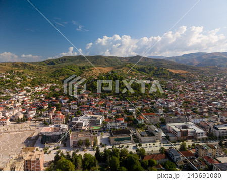 Berat, Albania spreads across valley beneath green hills, with dense homes and red tile roofs. Aerial town panorama includes broad plaza, tree lined streets, distant mountains and bright cloud Berat, Albania spreads across valley beneath green hills, with dense homes and red tile roofs. Aerial town panorama includes broad plaza, tree lined streets, distant mountains and bright cloud 143691080