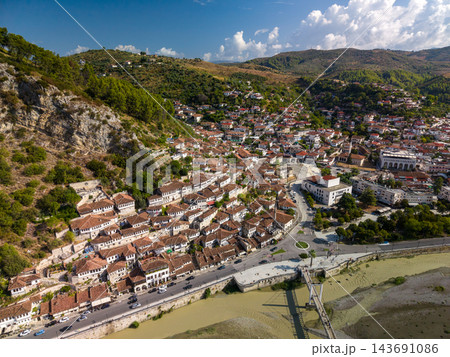 Berat Albania spreads across hillside beneath bright clouds, with traditional white houses and red tiled roofs. Historic townscape follows riverfront road beside green hills and stone riverbanks. Berat Albania spreads across hillside beneath bright clouds, with traditional white houses and red tiled roofs. Historic townscape follows riverfront road beside green hills and stone riverbanks. 143691086