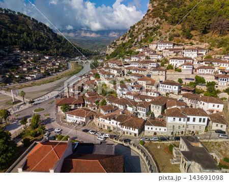 Berat Albania fills mountain valley with tightly clustered white houses, stone walls and red tiled roofs. Historic riverside town centers on arched bridge beneath dramatic clouds and green hills. Berat Albania fills mountain valley with tightly clustered white houses, stone walls and red tiled roofs. Historic riverside town centers on arched bridge beneath dramatic clouds and green hills. 143691098