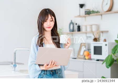 家のキッチンでコーヒーを片手にノートパソコンを見る笑顔の若い女性。くつろぎながら楽しむ日常生活 家のキッチンでコーヒーを片手にノートパソコンを見る笑顔の若い女性。くつろぎながら楽しむ日常生活 143691344