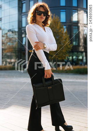 Beautiful Businesswoman Standing In City Holding Black Bag Wearing Stylish Outfit And Sunglasses. Corporate Fashion Lifestyle Concept Female Professional Urban Portrait Beautiful Businesswoman Standing In City Holding Black Bag Wearing Stylish Outfit And Sunglasses. Corporate Fashion Lifestyle Concept Female Professional Urban Portrait 143693065