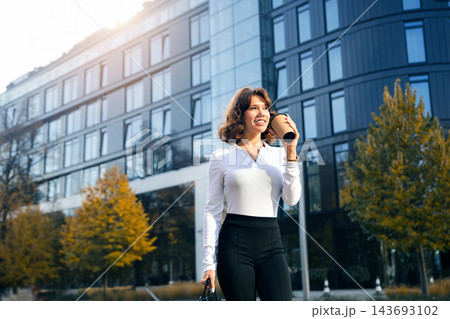 Beautiful woman drinking coffee while walking in business district. Attractive woman carrying office briefcase beside modern architecture. Corporate lifestyle concept. 143693102