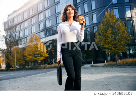 Young woman walking with takeaway coffee in elegant office outfit. Beautiful woman holding business briefcase near corporate headquarters. Professional success concept. 143693103