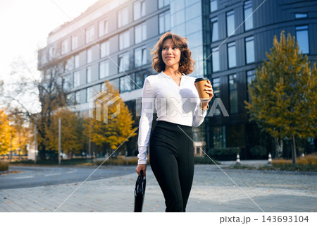 Attractive woman carrying coffee and business bag with confidence. Pretty woman wearing elegant office clothing near modern business center. Executive lifestyle concept. 143693104