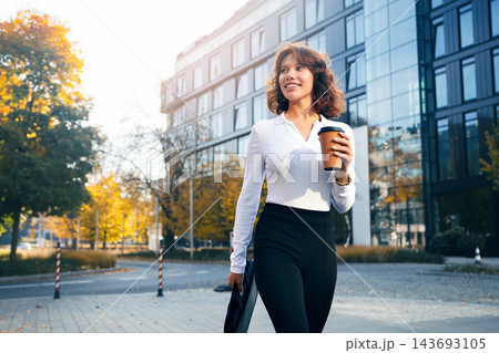 Pretty woman smiling with coffee outside contemporary office building. Attractive woman holding business briefcase in elegant professional style. Urban career concept. 143693105