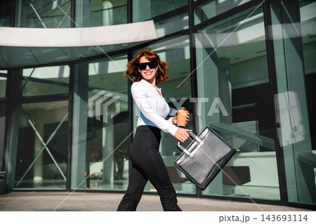 Beautiful woman carrying business briefcase with takeaway coffee outdoors. Pretty woman walking near contemporary office architecture confidently. Career growth concept. 143693114