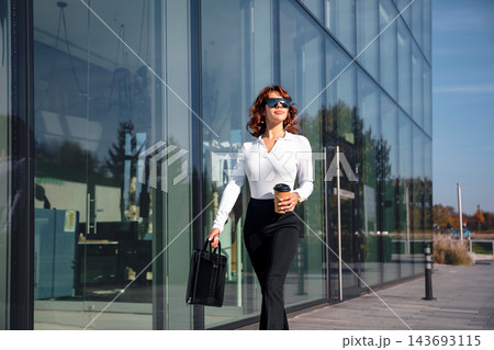 Beautiful woman walking with takeaway coffee beside modern glass office. Attractive woman carrying elegant briefcase in stylish business attire. Corporate lifestyle concept. Beautiful woman walking with takeaway coffee beside modern glass office. Attractive woman carrying elegant briefcase in stylish business attire. Corporate lifestyle concept. 143693115