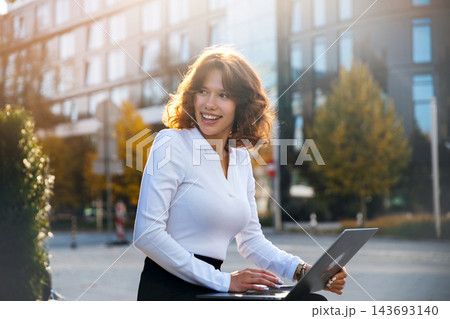 Beautiful woman working on laptop with confident professional smile. Young woman enjoying flexible business lifestyle outdoors. Remote office concept. 143693140