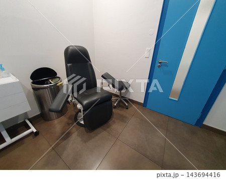 Medical examination chair with adjustable arm rests positioned in a clean clinic room next to a blue door and waste bin, suggesting healthcare, patient treatment and clinical procedures. 143694416