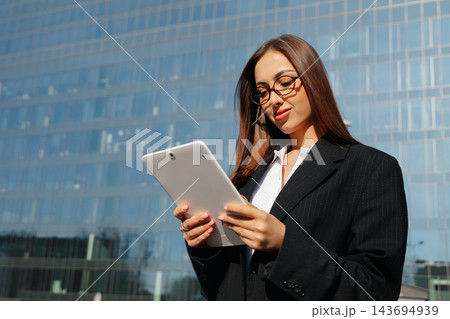 Attractive Business Woman Holding Tablet And Looking Down Near Modern Office Building Wearing Black Blazer. Corporate Lifestyle Concept Female Professional 143694939