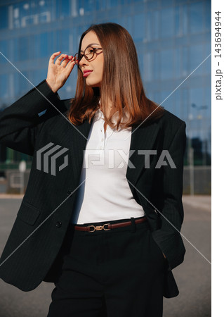 Attractive Business Woman Adjusting Glasses Near Modern Office Building Wearing Black Suit And White Shirt. Corporate Lifestyle Concept Female Professional Attractive Business Woman Adjusting Glasses Near Modern Office Building Wearing Black Suit And White Shirt. Corporate Lifestyle Concept Female Professional 143694944