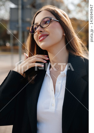 Attractive Business Woman Touching Neck And Looking Aside Near Modern Office Building Wearing Black Suit And Glasses. Corporate Lifestyle Concept Female Professional 143694947