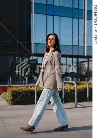 Attractive Woman Walking In City Wearing Beige Blazer And Jeans Near Modern Office Building. Casual Business Lifestyle Concept Female Urban Style Attractive Woman Walking In City Wearing Beige Blazer And Jeans Near Modern Office Building. Casual Business Lifestyle Concept Female Urban Style 143694961