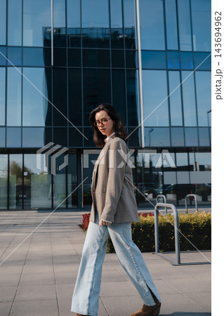 Pretty Woman Walking Near Glass Office Building Wearing Beige Blazer And Jeans. Casual Business Lifestyle Concept Female Urban Fashion Style 143694962