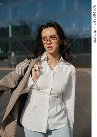 Pretty Woman In White Shirt And Glasses Holding Jacket Near Modern Office Building. Casual Business Lifestyle Concept Female Urban Portrait 143694976