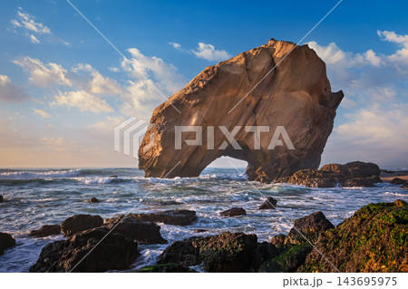 Penedo do Guincho stands majestically at Praia da Santa Cruz, framed by ocean waves and soft sands in warm late afternoon light. 143695975