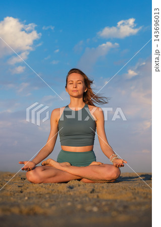 Facing forward on the beach, a woman holds Padmasana yoga, Lotus Pose, with closed eyes and upturned palms resting on her knees under a blue sky 143696013