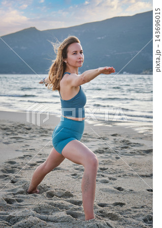 A woman holds Virabhadrasana 2 yoga, Warrior 2 Pose, bending her front knee and extending both arms level in a three-quarter beach view near a coastal hill 143696015