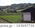 シャッターの閉まった倉庫と田んぼと緑の木々の森と山と雲の多い空の風景 143697736