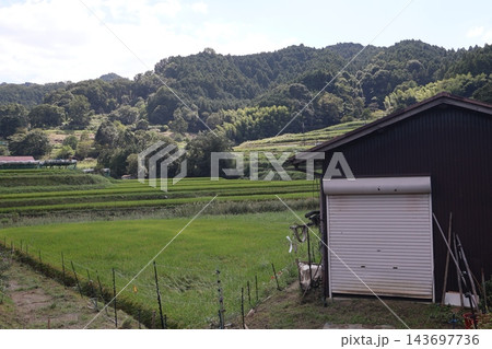 シャッターの閉まった倉庫と田んぼと緑の木々の森と山と雲の多い空の風景 シャッターの閉まった倉庫と田んぼと緑の木々の森と山と雲の多い空の風景 143697736