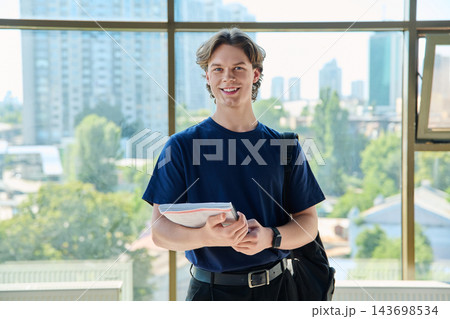 Young male student holding copybook standing looking at camera near window in campus 143698534