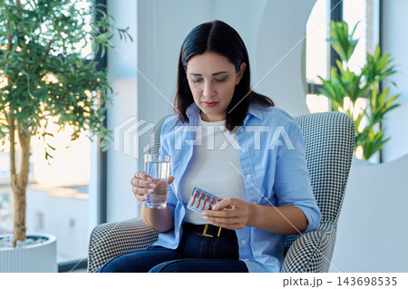 Woman holding glass of water and pills pack sitting in armchair 143698535