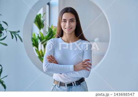 Smiling young female student with crossed arms standing in modern office or campus setting Smiling young female student with crossed arms standing in modern office or campus setting 143698547