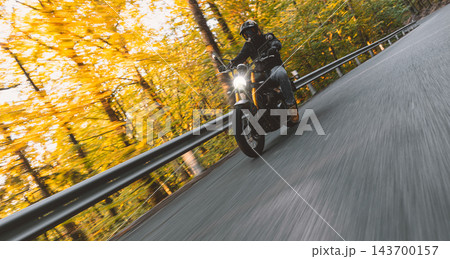 Motorcyclist wearing a black helmet and dark riding gear approaching the camera on a winding asphalt road through a sunlit green and golden autumn forest, with dynamic motion blur. Motorcyclist wearing a black helmet and dark riding gear approaching the camera on a winding asphalt road through a sunlit green and golden autumn forest, with dynamic motion blur. 143700157