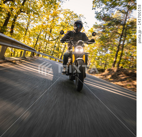 Motorcyclist wearing a black helmet and dark riding gear approaching the camera on a winding asphalt road through a sunlit green and golden autumn forest, with dynamic motion blur. Motorcyclist wearing a black helmet and dark riding gear approaching the camera on a winding asphalt road through a sunlit green and golden autumn forest, with dynamic motion blur. 143700158