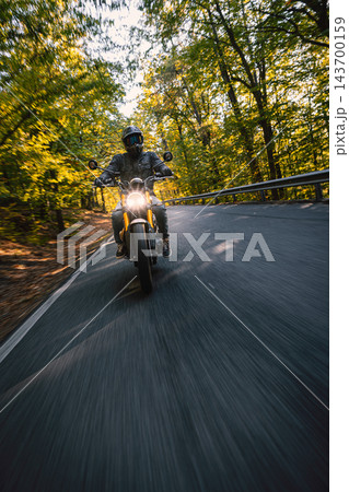 Motorcyclist wearing a black helmet and dark riding gear approaching the camera on a winding asphalt road through a sunlit green and golden autumn forest, with dynamic motion blur. Motorcyclist wearing a black helmet and dark riding gear approaching the camera on a winding asphalt road through a sunlit green and golden autumn forest, with dynamic motion blur. 143700159