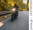 Motorcyclist wearing a black helmet and dark riding gear approaching the camera on a winding asphalt road through a sunlit green and golden autumn forest, with dynamic motion blur. 143700160