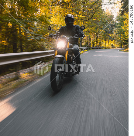 Motorcyclist wearing a black helmet and dark riding gear approaching the camera on a winding asphalt road through a sunlit green and golden autumn forest, with dynamic motion blur. Motorcyclist wearing a black helmet and dark riding gear approaching the camera on a winding asphalt road through a sunlit green and golden autumn forest, with dynamic motion blur. 143700160